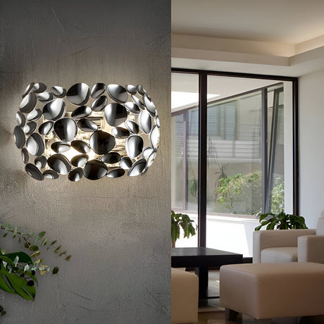 Contemporary living room with a wall sconce featuring a cluster of reflective, circular metal pieces. The room has large windows, white sofas, a black coffee table, and potted plants.