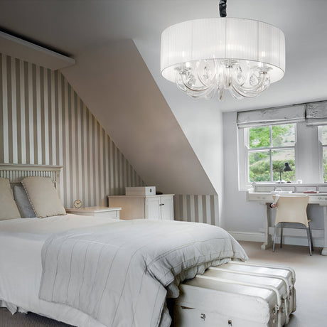 A cozy bedroom with a sloped ceiling, featuring a striped accent wall, white bedding, a white dresser, a large round chandelier, and a desk by a window with green foliage visible outside.