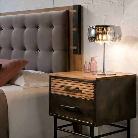 A modern bedside setup with a tufted headboard, a wooden nightstand featuring metal accents, a glass lamp, and pink decorative bottles. The bedding includes white sheets and a pink pillow.