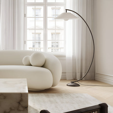 Minimalist living room with a curved white sofa, two round cushions, a tall arched floor lamp, and large windows with sheer curtains. The decor includes a marble coffee table and light wood flooring.