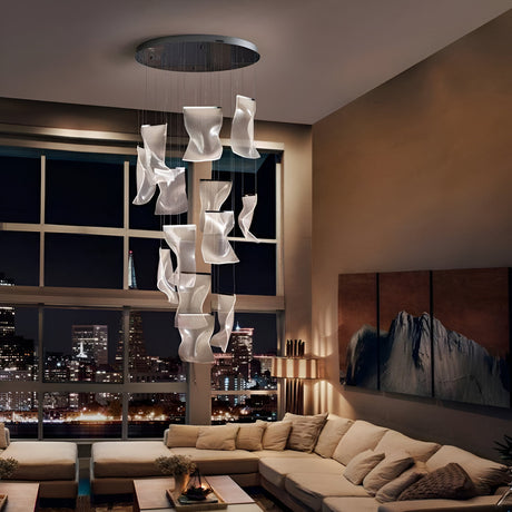 Modern living room with a large, abstract chandelier made of white, flowing shapes hanging from the ceiling. Beige sectional sofa surrounds a rectangular coffee table. Large windows offer a cityscape view.