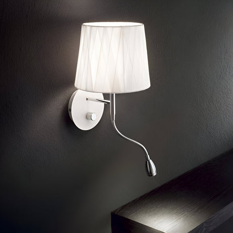 Wall-mounted lamp with a white pleated lampshade and a chrome base against a dark wall. A small, flexible reading light extends below, illuminating the surface beneath.