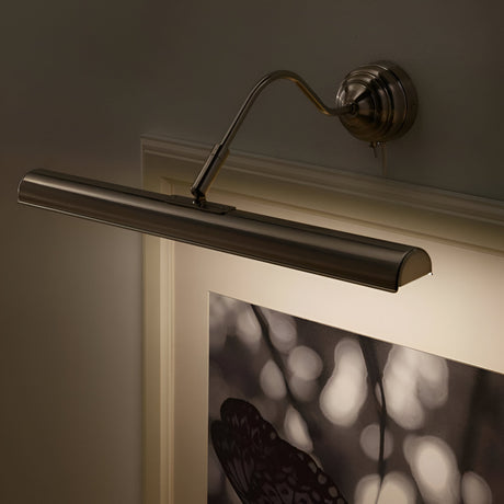 A wall-mounted, brushed metal picture light illuminates an art piece below. The artwork features an abstract design with blurred shapes. The light has an elegantly curved arm for focused display lighting.