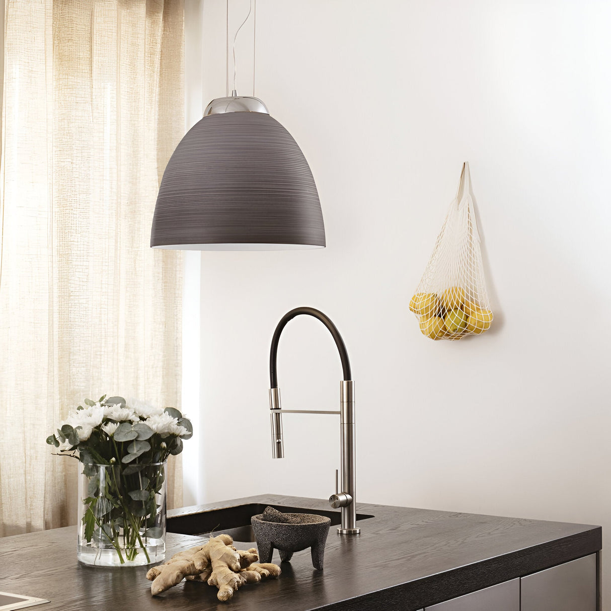 A modern kitchen with a dark countertop showcasing a vase of white flowers, a mortar and pestle, and ginger. A sleek faucet is centered under the striking Archer 1 Light Pendant in grey. Lemons hang in a net on the white wall, complemented by nearby beige curtains.