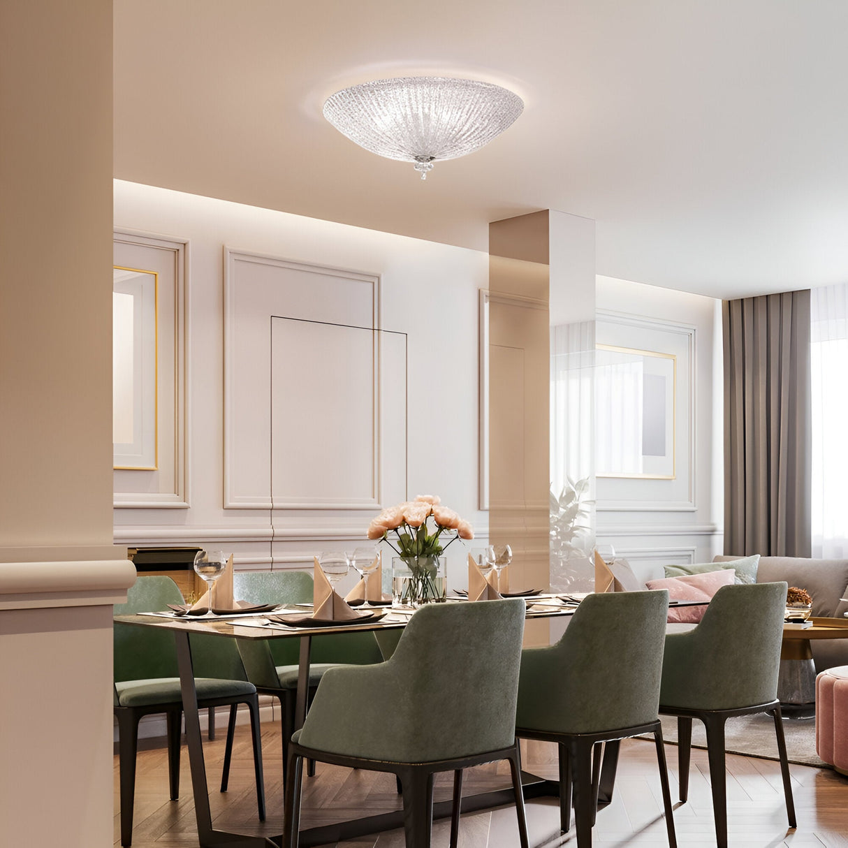 A modern dining room showcases a set table with green velvet chairs, highlighted by chrome metal accents. A bouquet of pink flowers is centered beneath the Sapphire 4 Light Ceiling in Clear, complementing the space’s transparent glass elements. The room exudes elegance with stylish wall molding and soft lighting filtering through the curtains.