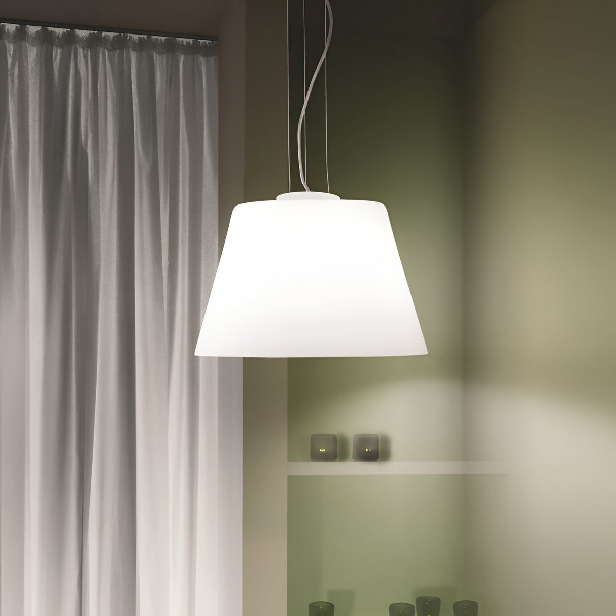 A sleek Haven 1 Light Pendant - White is suspended from the ceiling, gently illuminating a room with green walls. On the left, sheer white curtains cover a window, while small dark vases enhance the minimalist style of a shelf in the background.