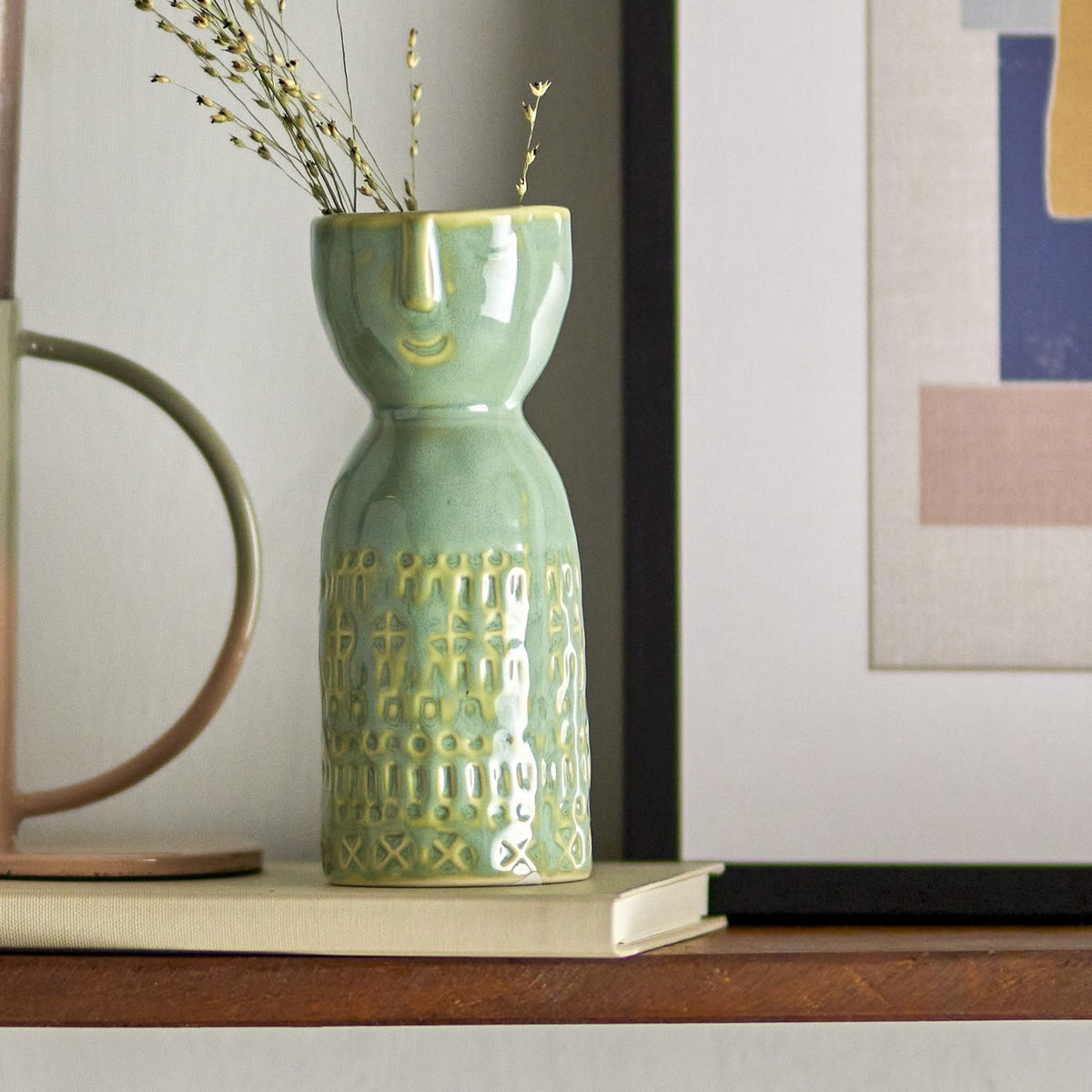 Embla Stoneware Vase 14.5cm in green ceramic with a sculpted face below the rim and a textured patterned base, shown on a shelf with a book, mug and dried stems.