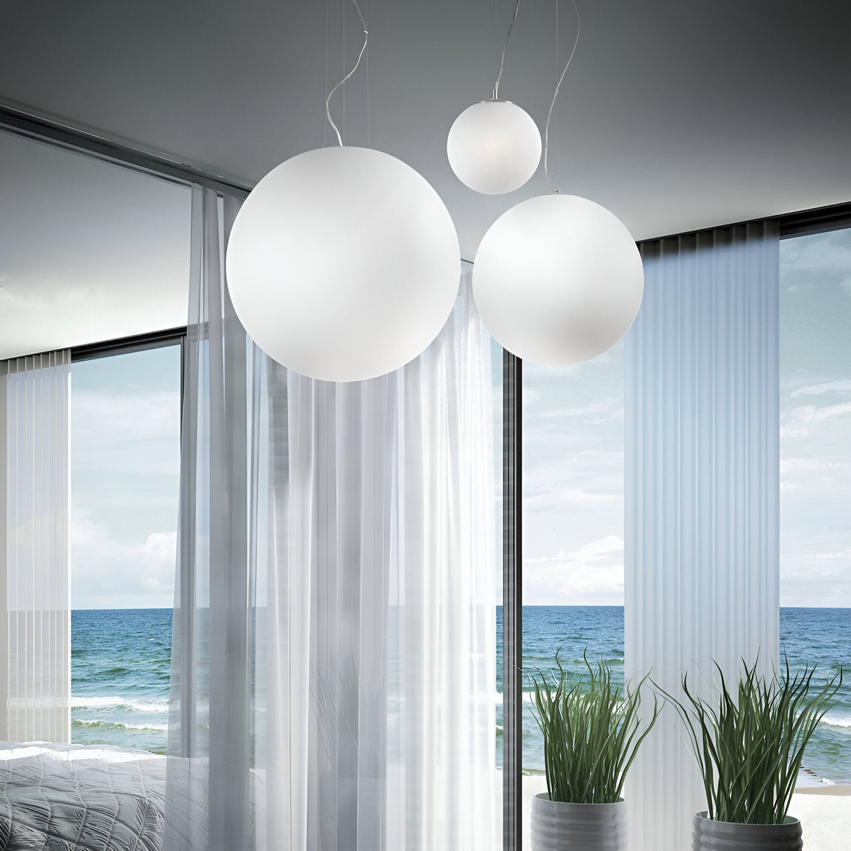 A modern room with large windows overlooking the ocean features three Lyria 1 Light Glass Pendants in white, each measuring 40cm, suspended from adjustable steel cables. Transparent curtains and potted plants add to the airy atmosphere of the space.