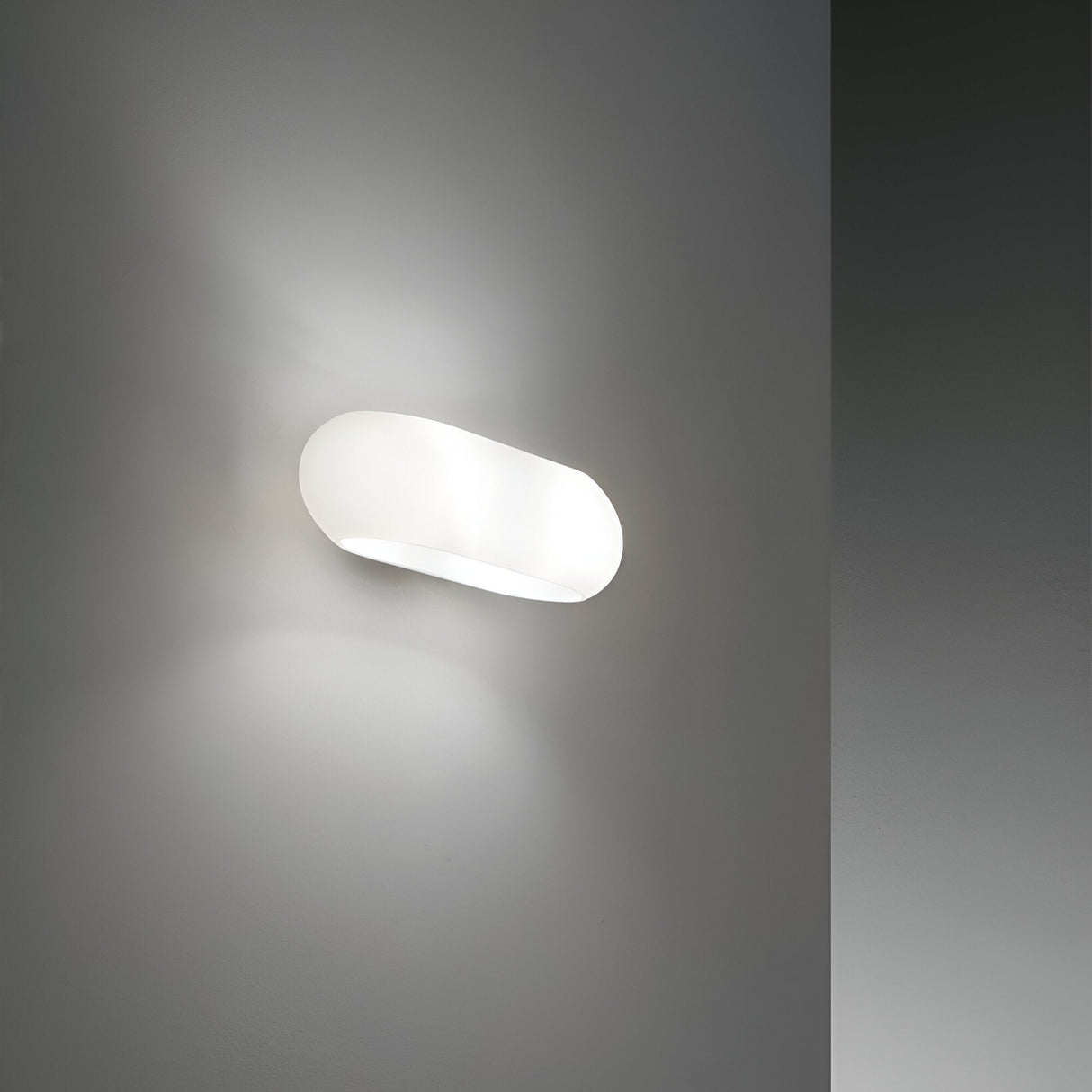 The Clip 2 Light Wall Light in white features a chrome frame that emits soft, diffused light against a grey wall, enhancing the minimalist decor and creating a calming ambiance. Its oval shape and sleek design are perfect for complementing modern interiors.