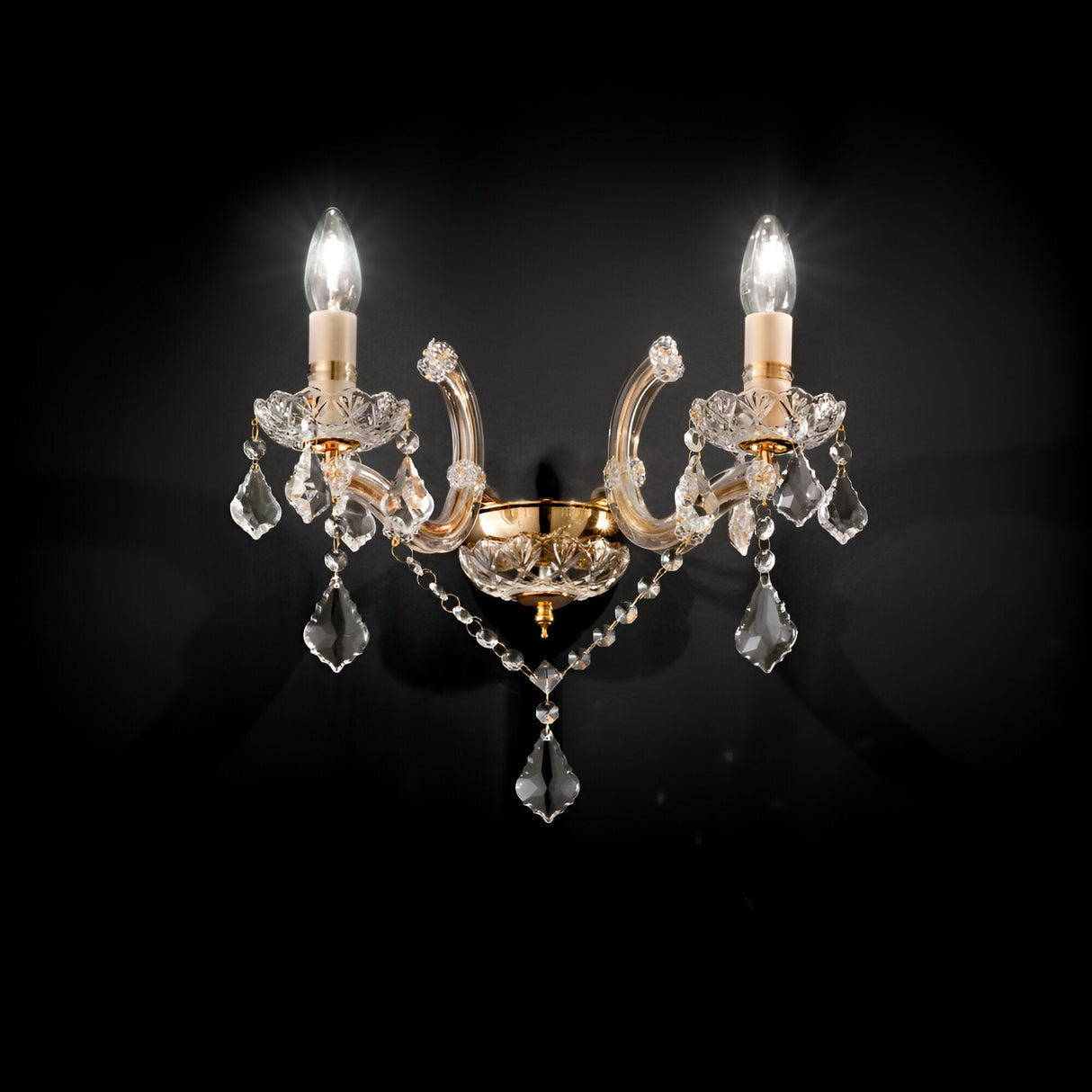 The Aurora Wall Crystal Light- Gold is a decorative sconce with two candle-shaped bulbs set on a gold and crystal base. It emits a soft glow, highlighting the intricate design embellished with crystal accents against a dark backdrop.
