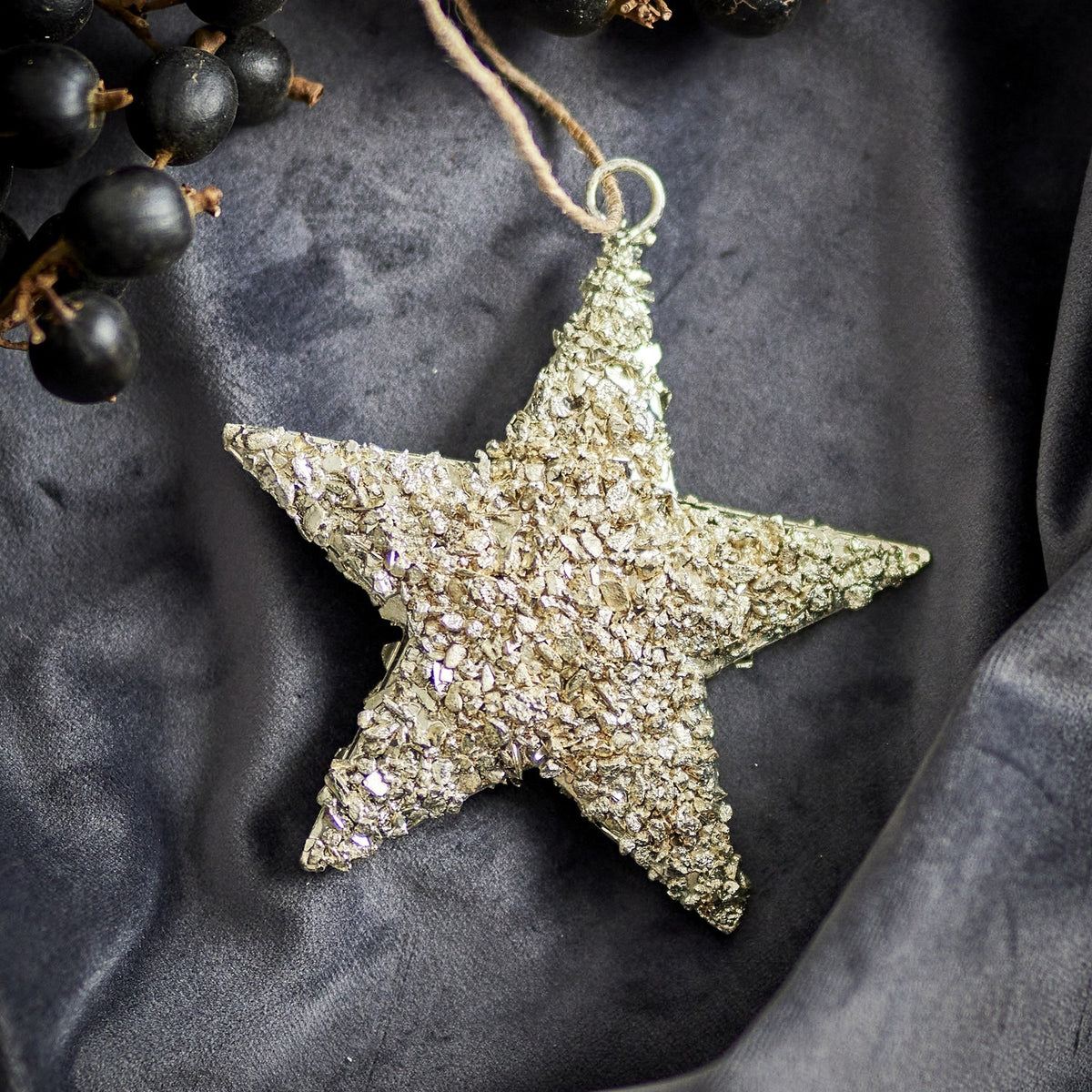 Sarika Star Iron Christmas Tree Decoration in gold with a textured, crumbly finish and jute string hanging loop, shown on a dark grey fabric backdrop with black berries.