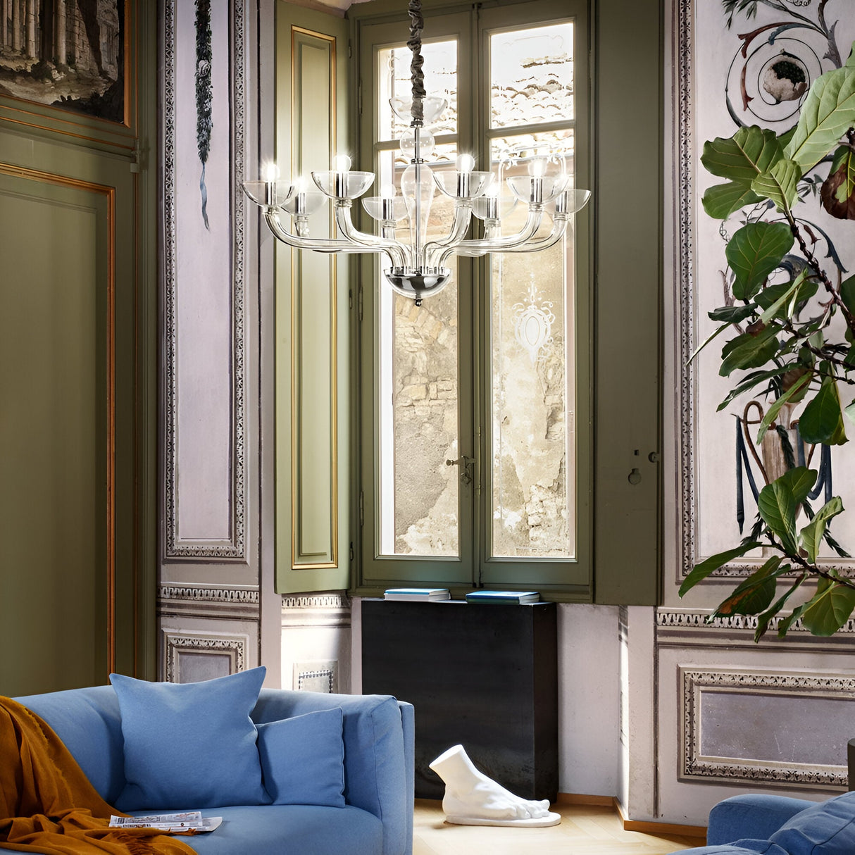 An elegant living room is highlighted by the Casio Glass 8 Light Chandelier in chrome, which hangs from the ceiling to illuminate a cozy blue sofa. The sophisticated ambiance is enhanced by a large window, classical moldings, and a decorative plant against intricately designed walls.