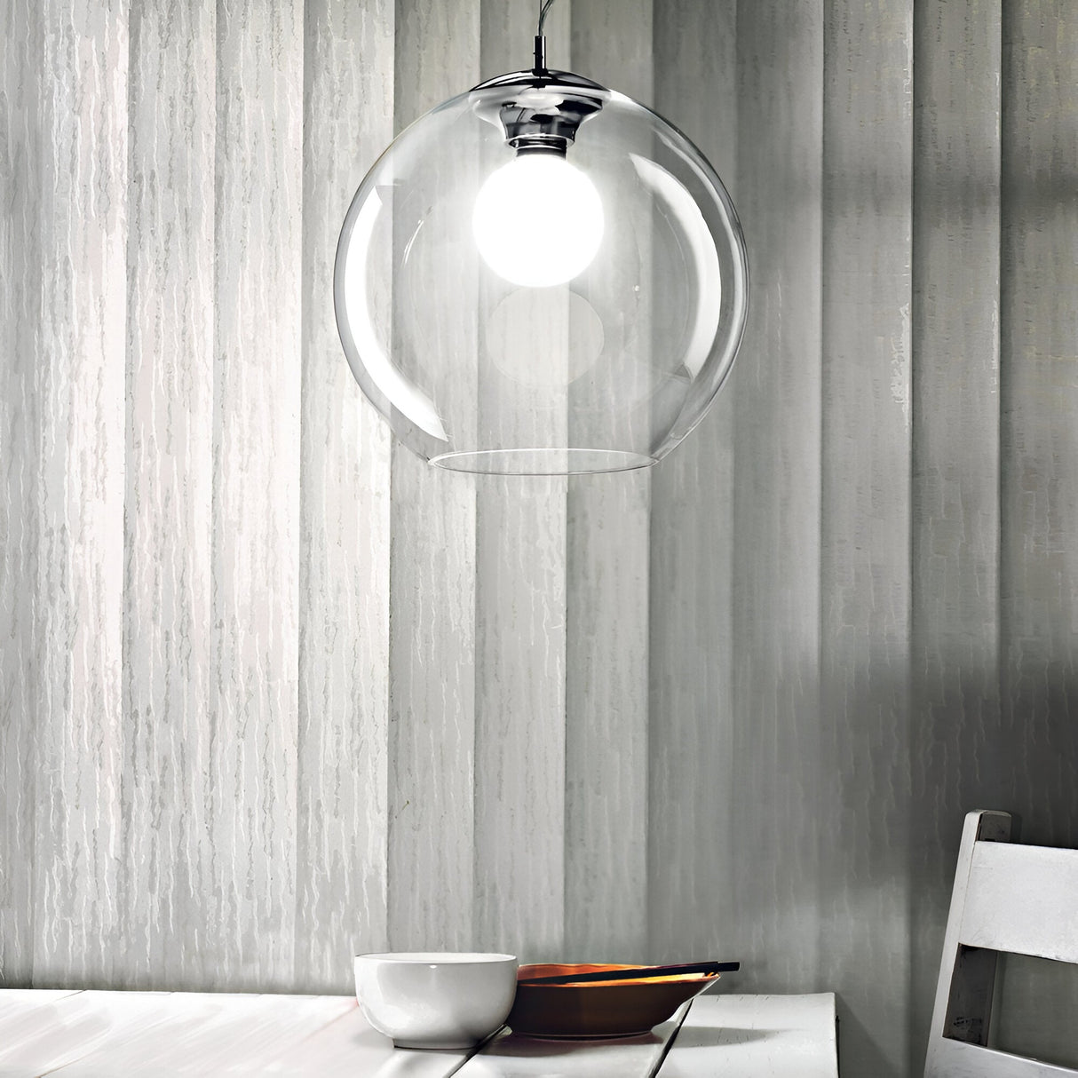 A modern dining area features a white table and bowl, illuminated by the Emberlyte 1 Light Glass Pendant in Clear, hanging overhead and offering bright, dimmable light. Textured grey walls form the backdrop, with a portion of a white chair visible on the right.