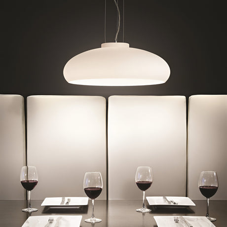 A modern dining setup features the Eclipsea 1 Light Pendant in white, elegantly illuminating a dark table. The arrangement includes four wine glasses filled with red wine and four square white plates, all set against a backdrop of tall, cushioned panels.