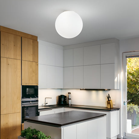 A modern kitchen featuring sleek white cabinets paired with a dark countertop. The tall cabinets are enhanced by wooden accents, perfectly complementing the black oven and coffee maker. Elegance is added by a large, dimmable Lyria 1 Light Flush in white, measuring 40cm, while a door opens to reveal lush garden greenery.