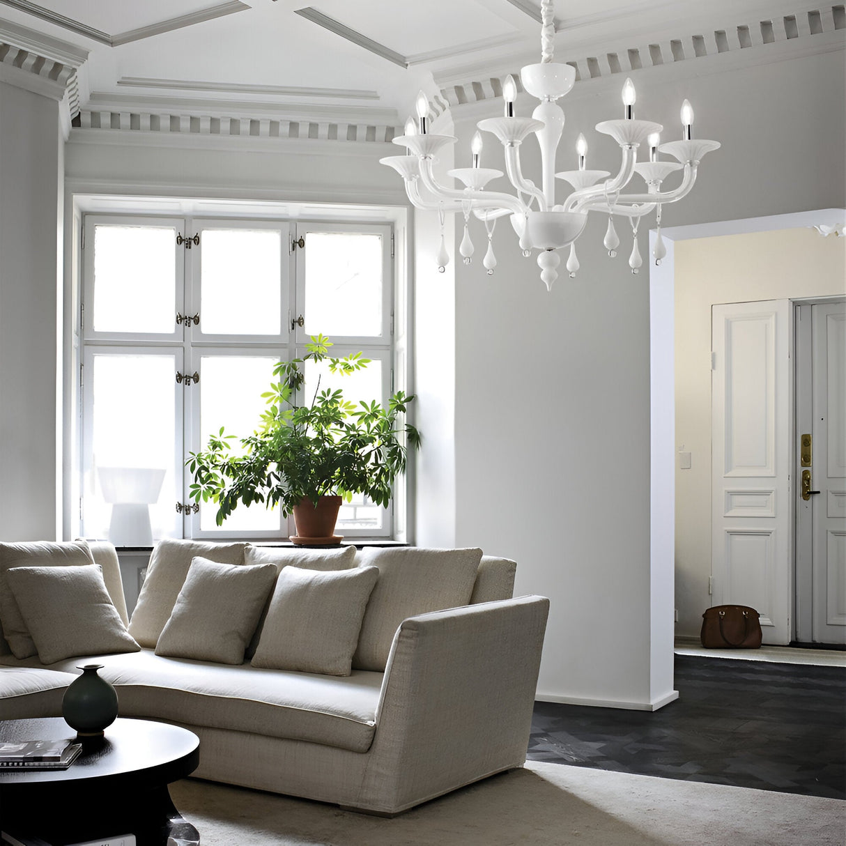 A modern living room showcasing a beige sectional sofa and a black round coffee table with a vase. The Nirvana 8 Glass Light Chandelier - White, with its adjustable chain length, elegantly graces the ceiling. Nearby, a potted plant flourishes by the window, and a white door is visible in the background.