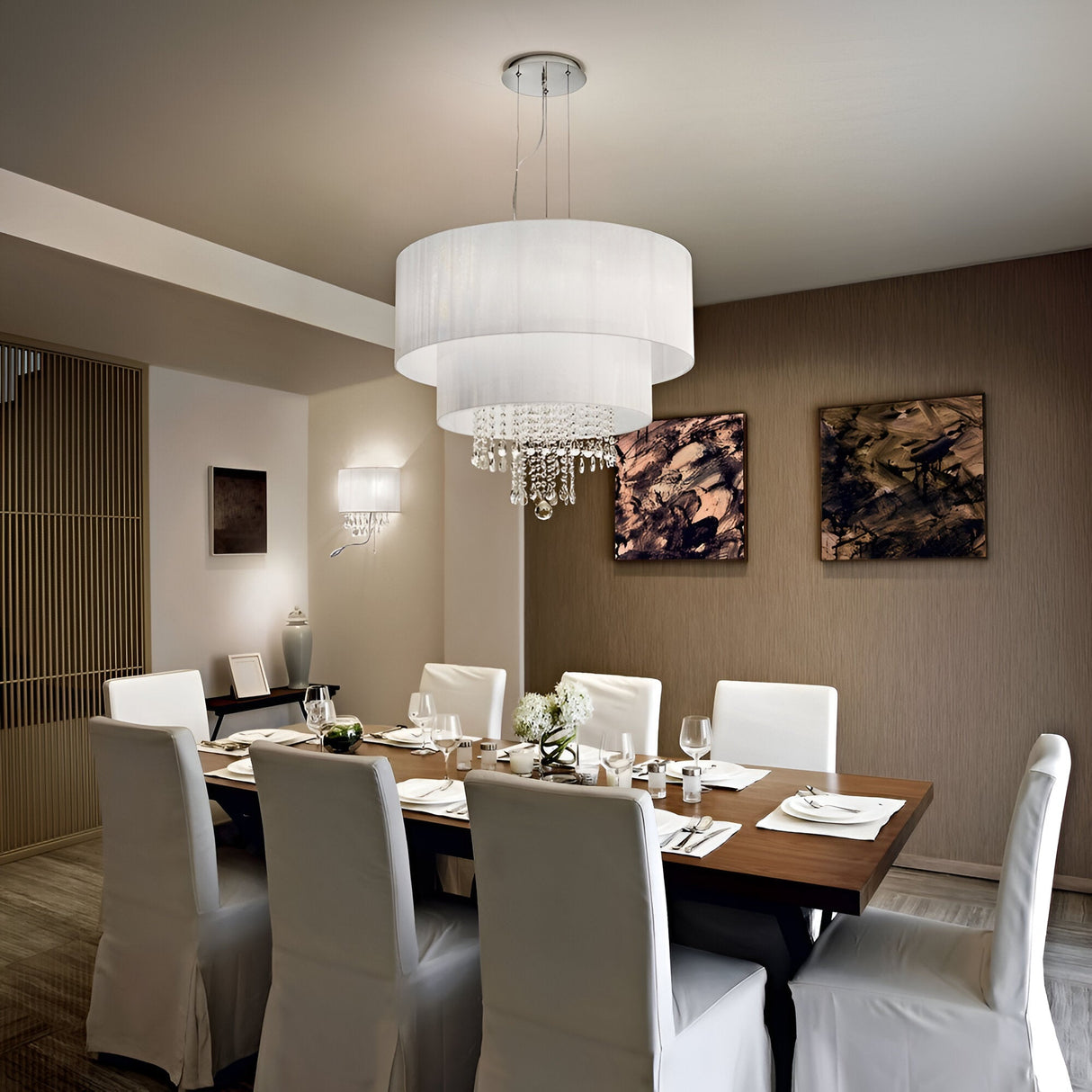 A contemporary dining room with a dark wooden table, eight white chairs, and the striking Brillis Crystal 6 Light Chandelier in white overhead. The walls feature abstract art pieces that complement the modern interior. The table is elegantly set with white tableware and a floral centerpiece.