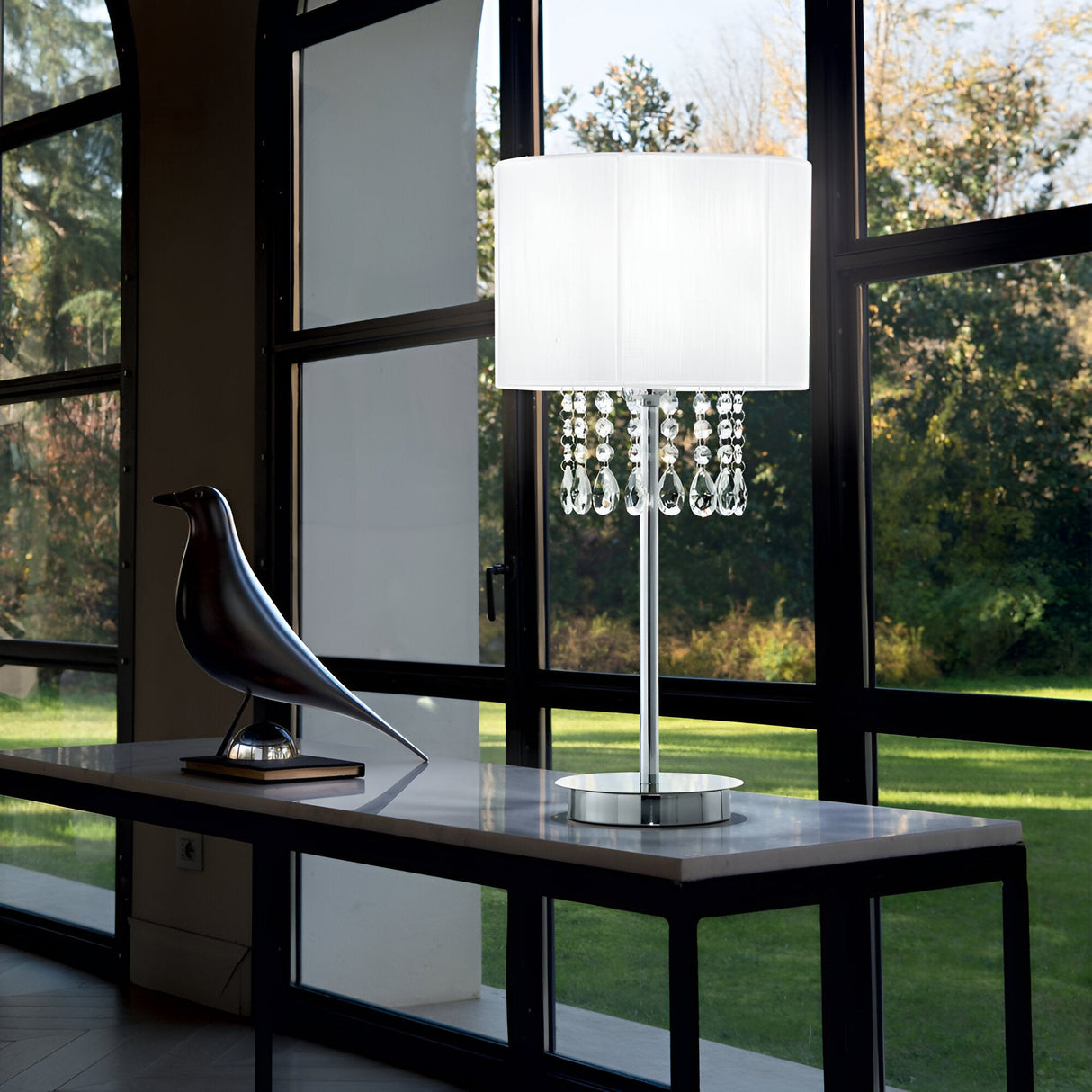 A contemporary indoor scene with a large window overlooking a lush green lawn exudes a serene atmosphere. A sleek table showcases a bird sculpture alongside the Brillis Crystal Table Lamp - White, its shade embellished with cascading crystals. Sunlight pours in, creating gentle shadows.