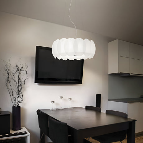 In a contemporary dining room, a dark wooden table is accompanied by four black chairs and an elegant Nero 5 Light Pendant in white with acid-etched glass. A large, circular pendant light with adjustable cables complements the flat-screen TV on the wall, alongside a minimalist kitchen area in the background.