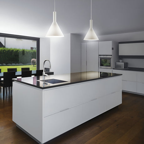 The minimalist kitchen showcases a large island equipped with a stovetop and sink. Above the island, two Vesper 1 Light Large Pendants in white emphasize modern lighting with their elegant design. In the dining area by the spacious windows, a table overlooks a garden adorned with green hedges. The dark wooden floor creates a striking contrast throughout the space.