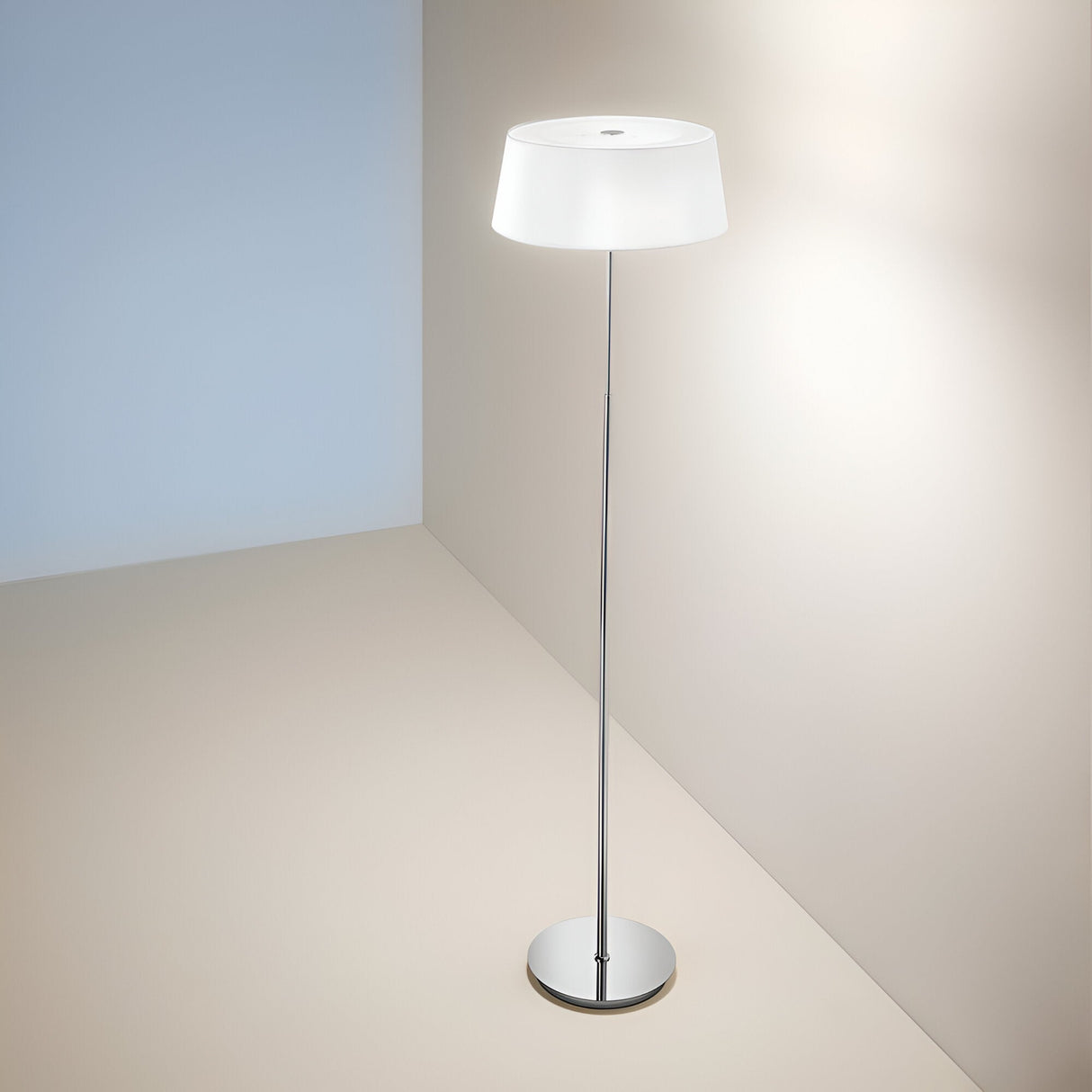 The Satin Floor Lamp - Silver/White, showcasing a modern design with its sleek silver stand and white lampshade, is positioned in the corner of a softly lit room featuring beige walls and a light-colored floor. Equipped with Bulb Type E14, the lamp emits a gentle glow that creates a cozy ambiance.
