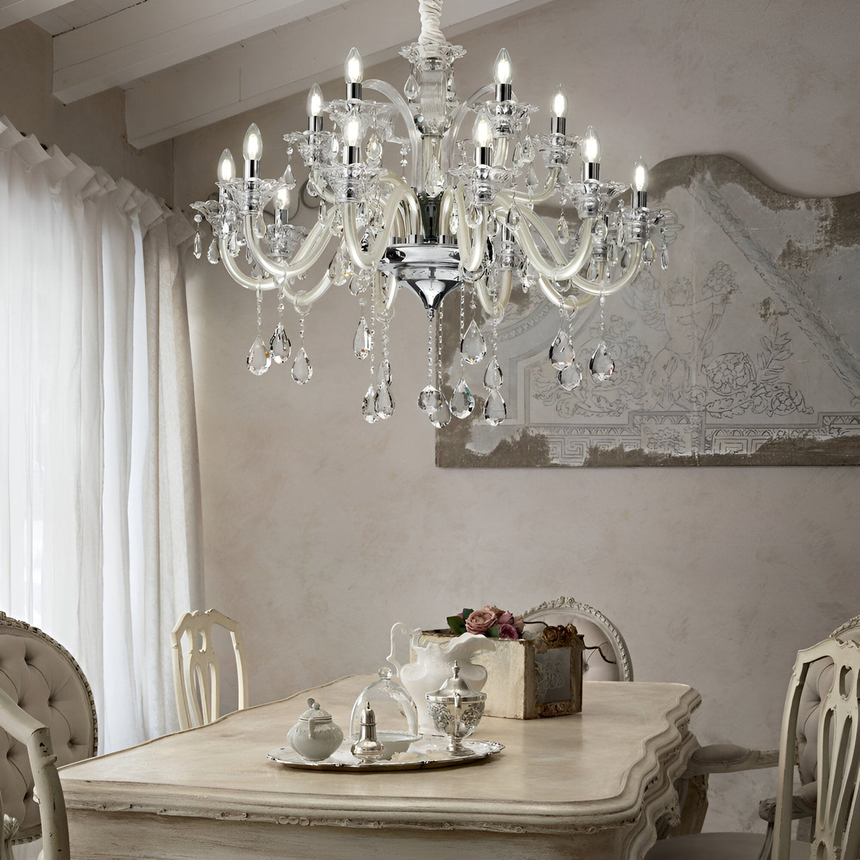 Elegant dining room with a vintage wooden table and upholstered chairs. A silver tea set is arranged on the table. Above, a Vivid Crystal 15 Light Chandelier - White with elegant lighting illuminates the space, enhancing its classic decor.