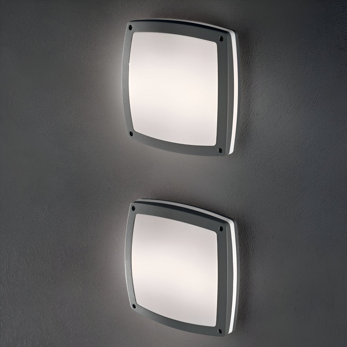 The Lyrisa 3 Light Flush Ceiling & Wall Light in Grey, with an IP54 safety rating, showcases two square modern fixtures with rounded edges on a grey wall. These bathroom lighting units come with dark frames and provide a soft, diffused glow for an elegant and minimalistic appearance.