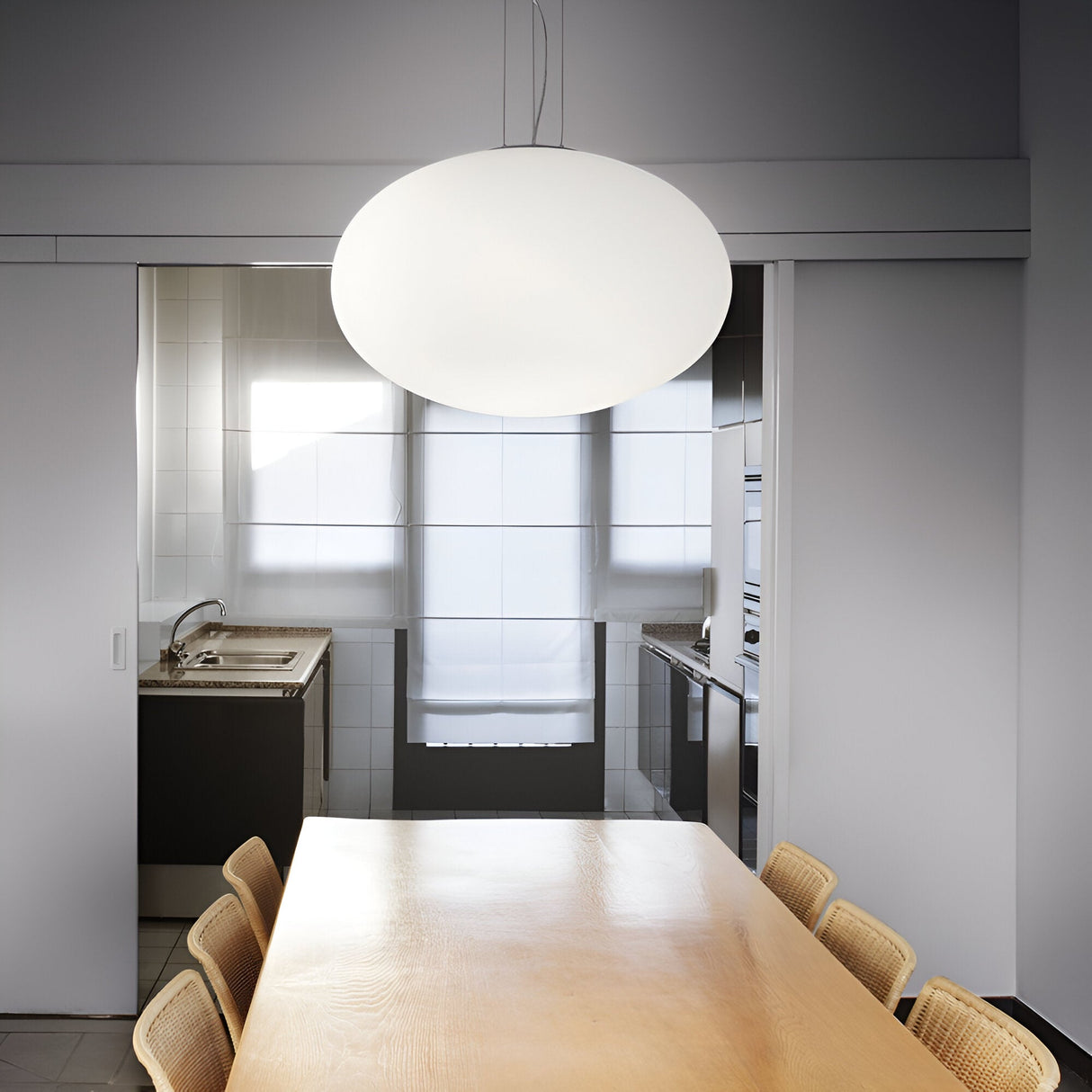 In a minimalist dining room, there's a large wooden table accompanied by six wicker chairs. Hanging above them is the 40cm white Look 1 Light Pendant, introducing a modern flair to the space. The background features a compact kitchen with tiled walls and a stainless steel sink, while natural light streaming through frosted windows enhances the contemporary interior design.