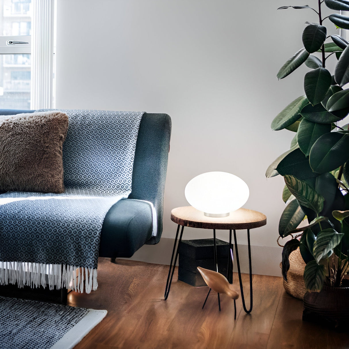 A modern living room showcases a patterned grey sofa, a fringed throw, and a brown cushion. A small round table holding the Look Table Lamp in a sleek white finish is positioned beside a large potted plant. The space exudes cozy minimalism with its warm wooden floor.