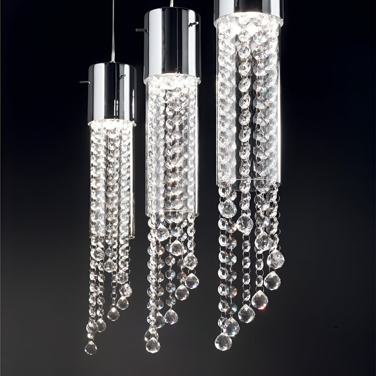 A trio of Cono 1 Light Glass Pendants in chrome, featuring sleek cylindrical designs and adorned with cascading crystal beads, illuminate a dark background with a gentle, luxurious glow.