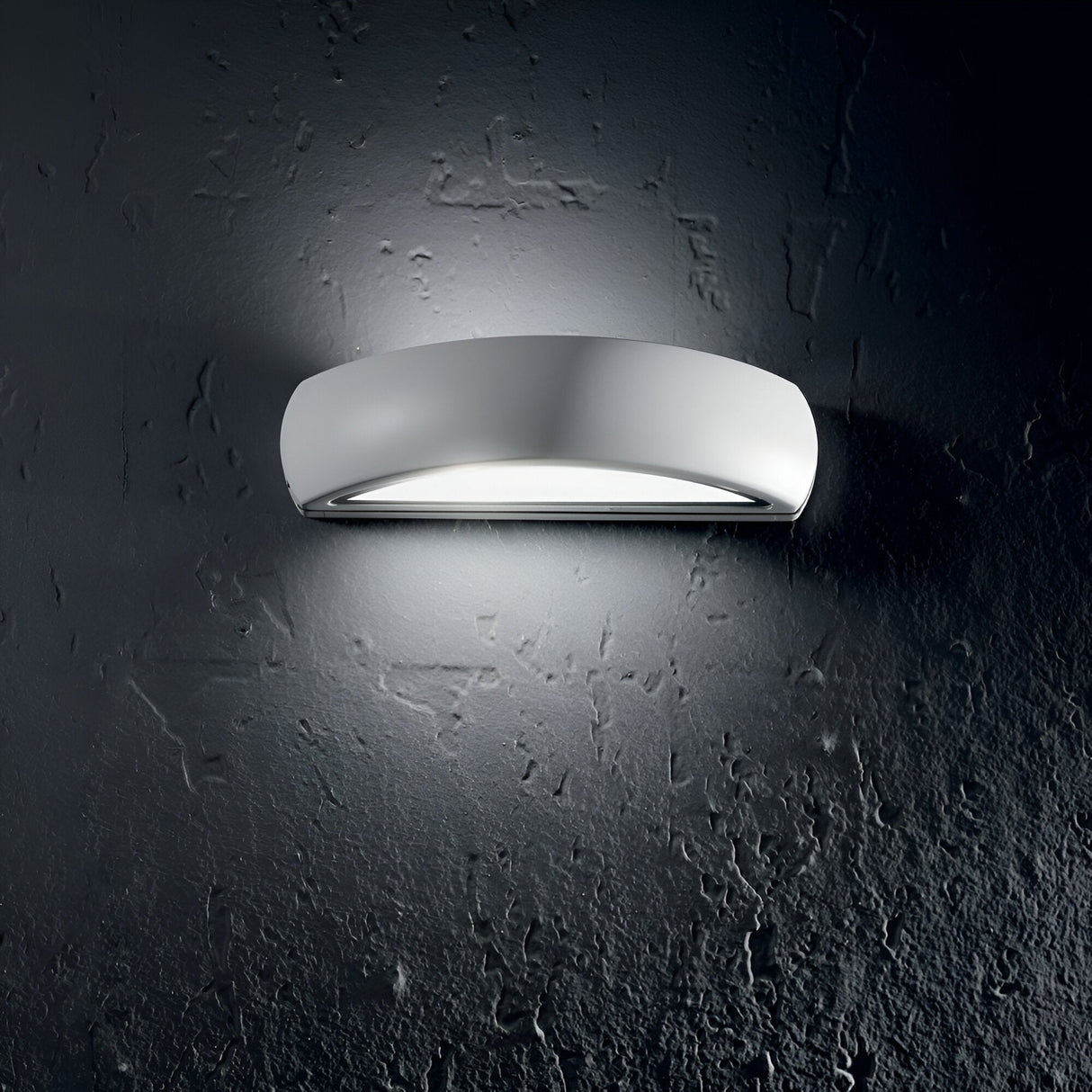 A modern Solara Outdoor Wall Light - White, made of powder-coated aluminum, illuminates a textured gray wall. The oval-shaped fixture, with its sleek minimalist design and IP54 rating, casts a soft glow onto the surface.