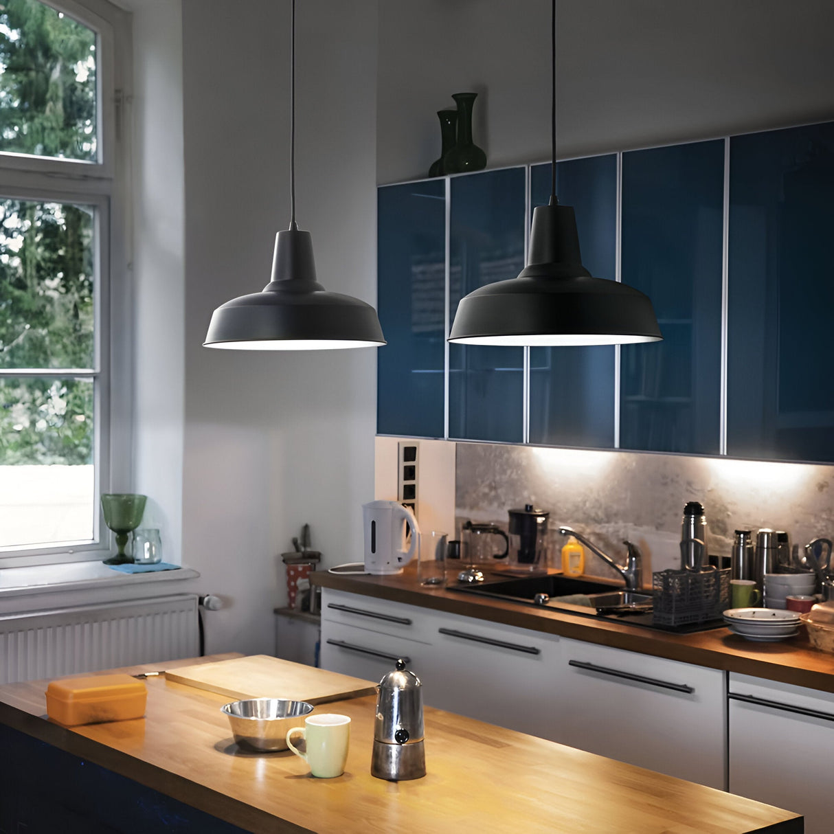 A modern kitchen features blue cabinets, a wooden countertop, and two Blaze 1 Light Pendants in black for an industrial touch. A coffee maker stands on the counter alongside mugs and bowls. The window reveals a green outdoor view, filling the space with natural light.