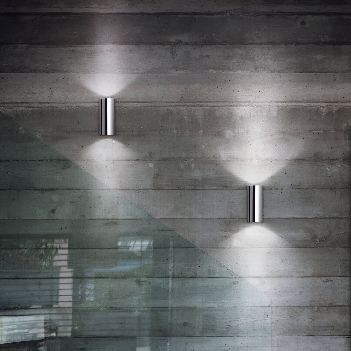 A modern interior showcases two Noctis 2 Light Wall Lights - Silver, mounted on a textured concrete wall and casting a soft glow upward and downward. In the foreground, a glass balustrade creates a diagonal line across the image, accentuating the contemporary design.