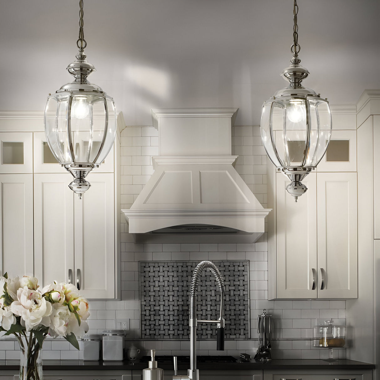 A modern kitchen exuding elegance showcases the Ulisse 1 Light Pendant - Chrome elegantly suspended from the ceiling. The space features white cabinetry, a stainless steel faucet, and a decorative tile backsplash. A vase with white flowers graces the counter, further enhancing its refined charm.