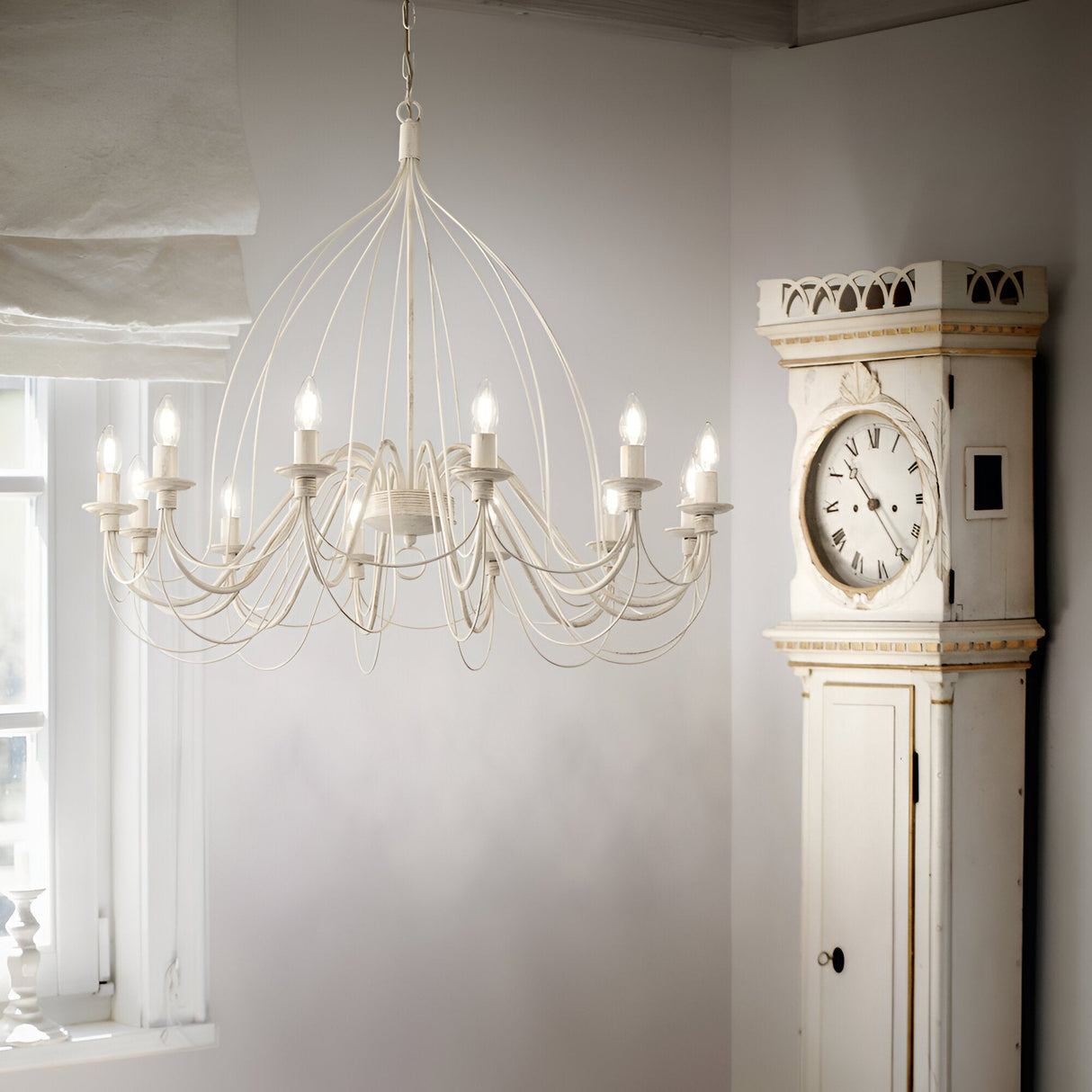 A Seraph 12 Light Chandelier with a hand-painted cream finish and multiple arms is suspended from the ceiling in a bright room. Beside it, a vintage white grandfather clock stands against the wall, near a partially open window with flowing white curtains.