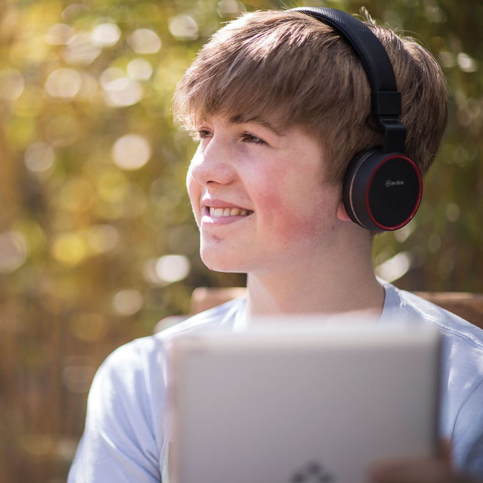 Young person wearing AVSL AV:Link PH10-BLK matt black headphones with red trim and cushioned earcups, smiling outdoors while using a tablet.