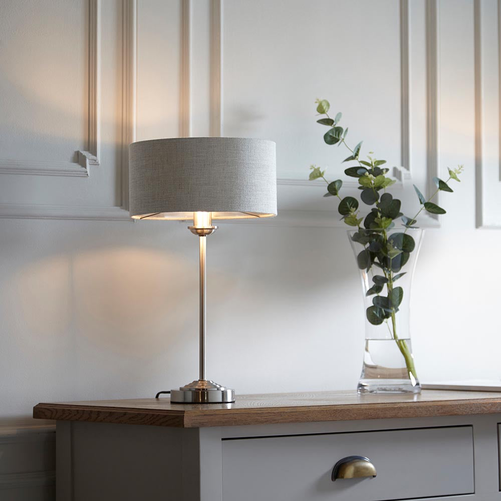 Brenley Table Lamp in brushed chrome with a natural linen drum shade, lit on a wooden sideboard beside a vase of eucalyptus.