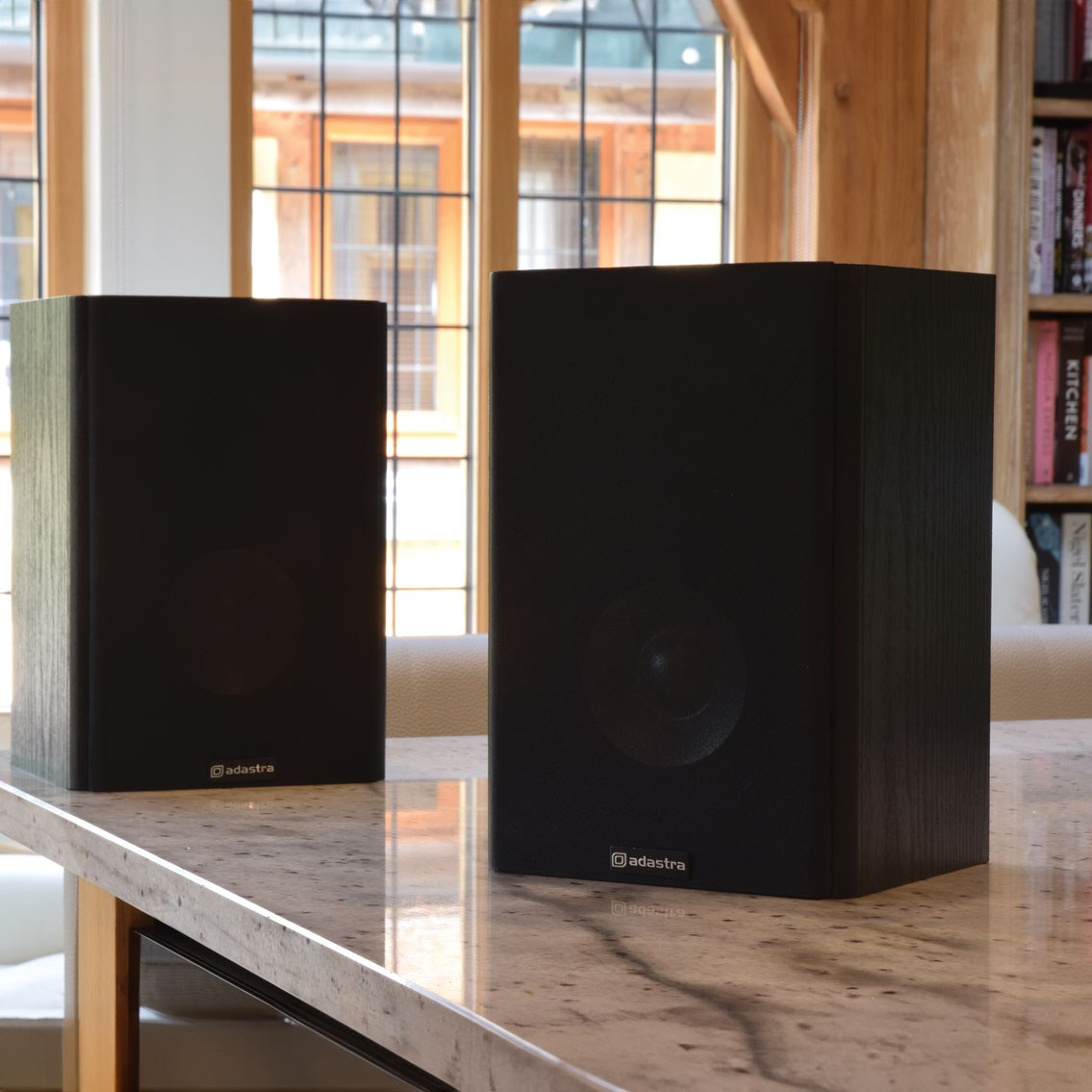 Adastra AB-5 passive bookshelf speakers in matt black woodgrain finish, displayed as a pair on a marble surface near a window.