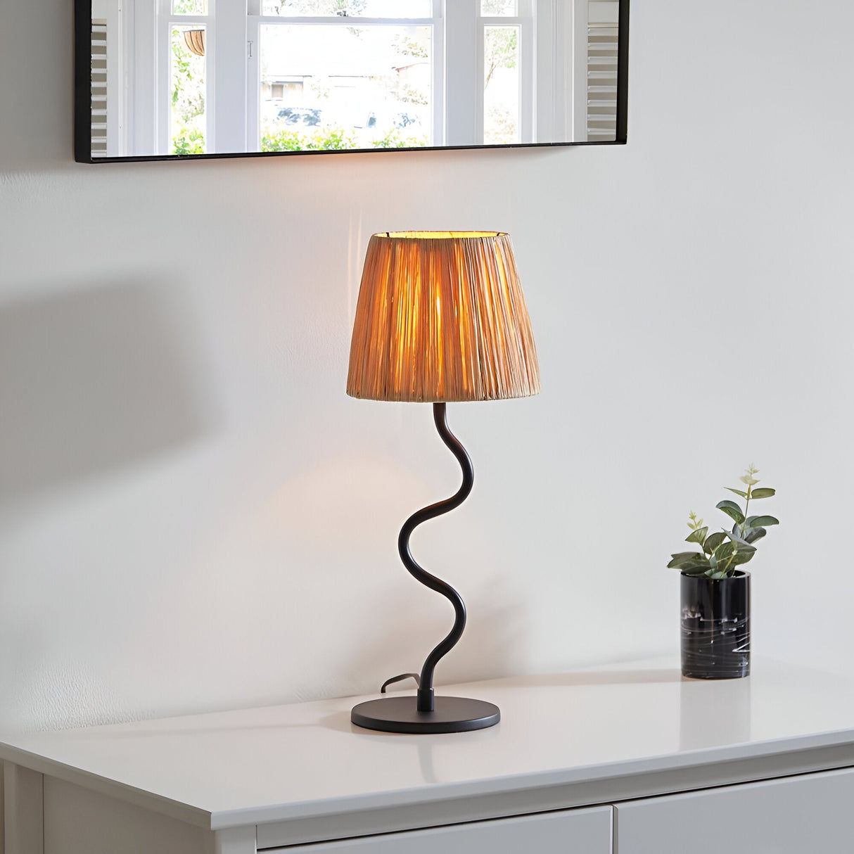 Endon Danemont table lamp with a wavy matt black metal stem and tapered natural raffia shade, on a white console beneath a black-framed mirror.