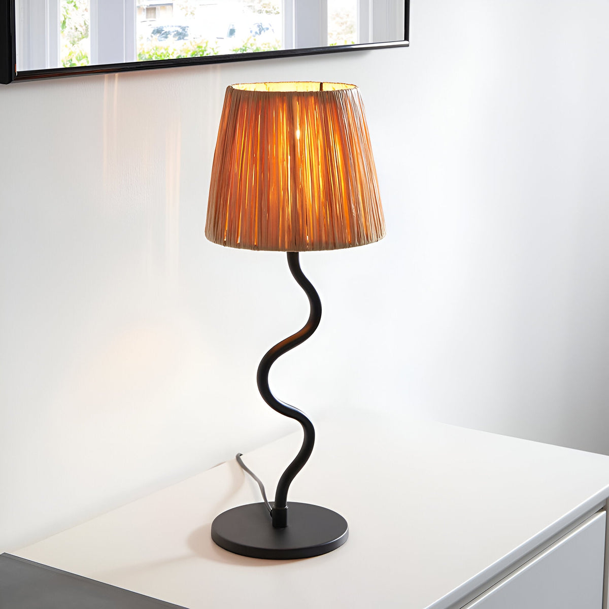 Endon Danemont table lamp with natural raffia tapered shade and wavy matt black metal stem on a round base, set on a white console beneath a mirror.