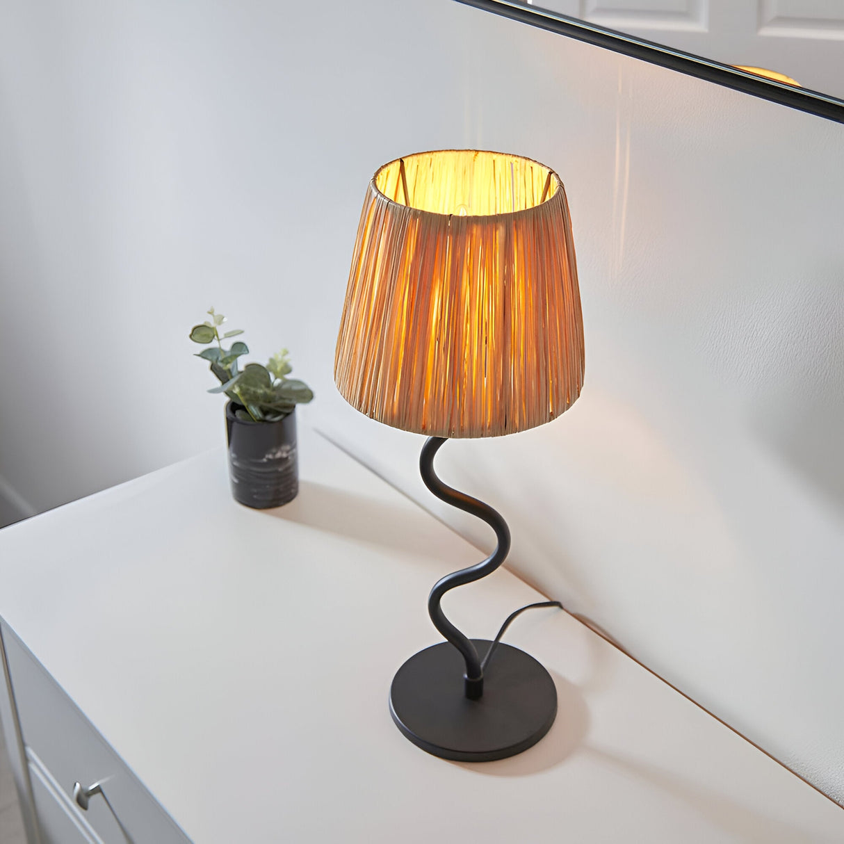 Endon Danemont table lamp with a natural raffia tapered shade on a wavy matt black metal stem and round base, lit on a white sideboard.