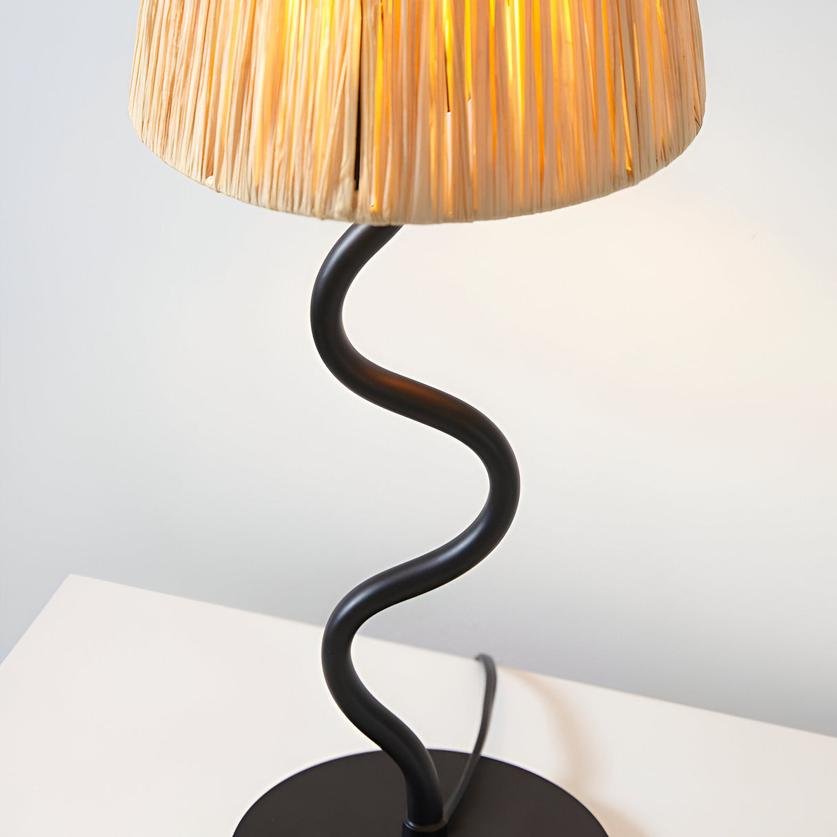 Endon Danemont table lamp with a natural raffia tapered shade and matt black wavy metal stem, illuminated on a white surface.