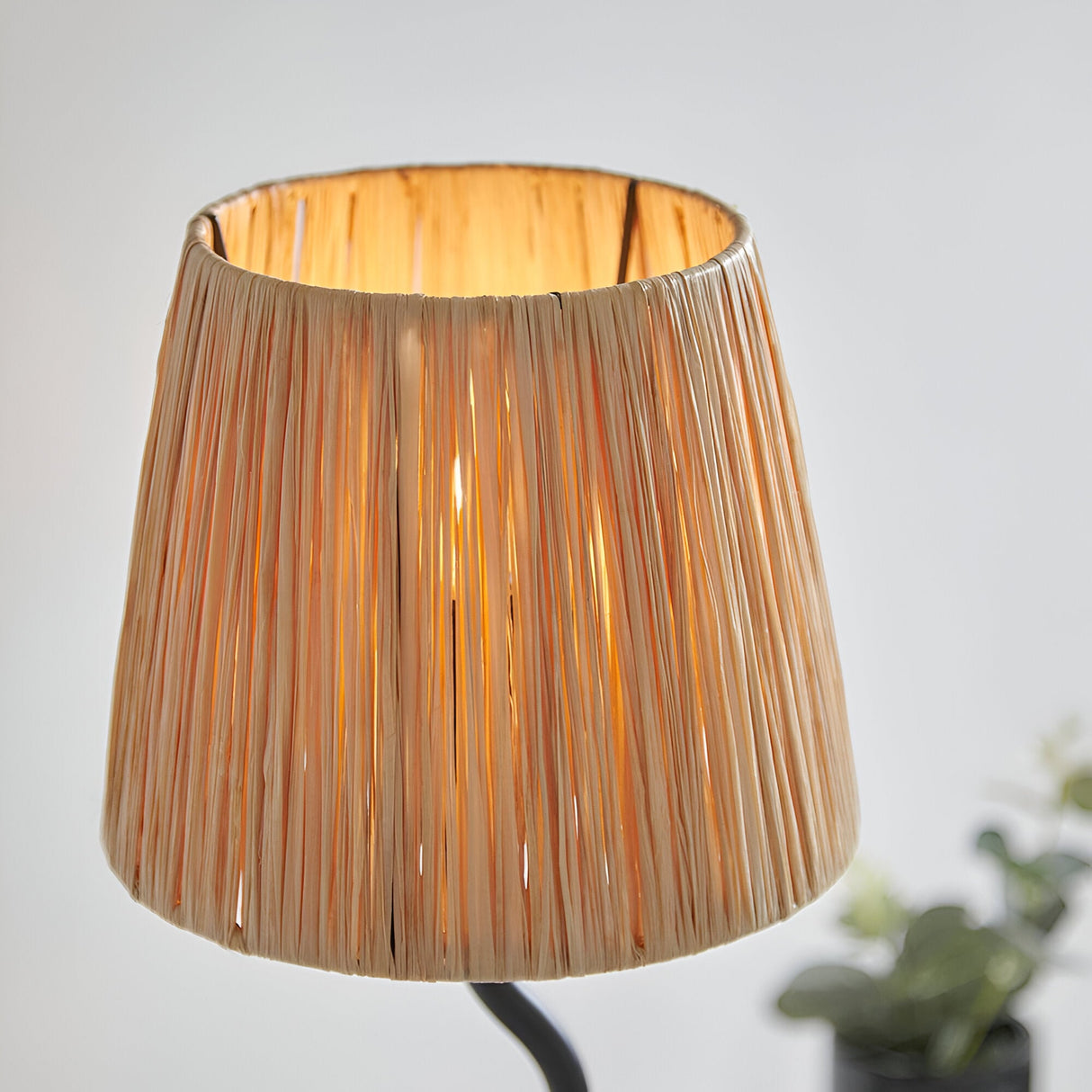 Endon Danemont table lamp with a tapered hand-wrapped natural raffia shade illuminated on a slim black metal frame, shown close-up indoors.