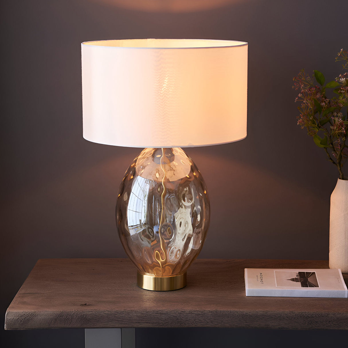 Endon Draycott table lamp with dimpled amber glass base, satin brass foot and white fabric drum shade, lit on a wooden console beside a vase.