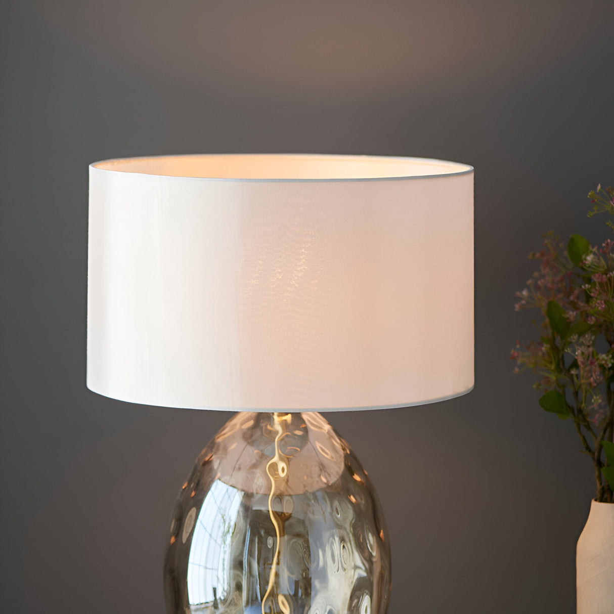 Endon Draycott table lamp with dimpled glass base and white fabric drum shade illuminated, shown against a dark grey wall beside pink blossoms.