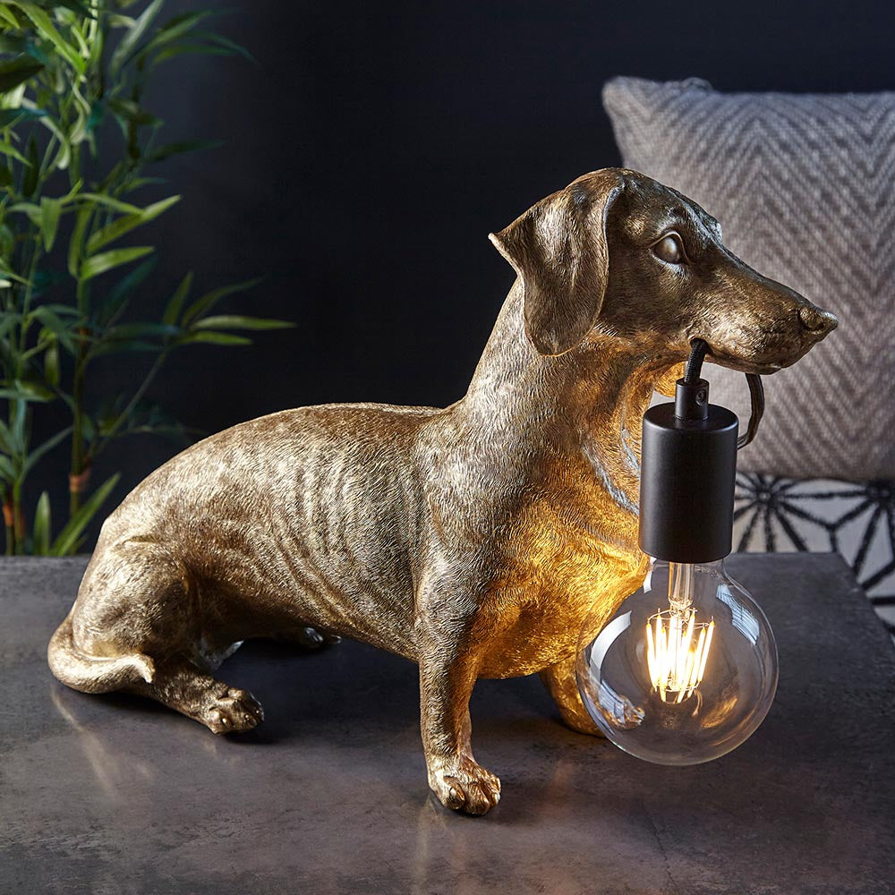Bevworth Table Lamp in gold resin dachshund design with black E27 bulb holder and lit filament globe bulb, displayed indoors near a plant and cushion.