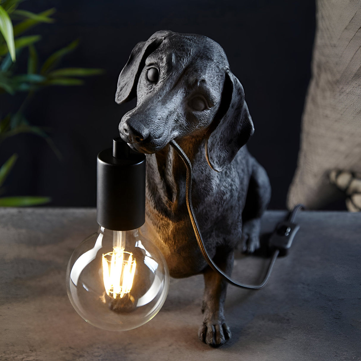 Bevworth Table Lamp in black resin, shaped as a dachshund holding an E27 filament bulb, matt black lamp holder, set against a dark background.
