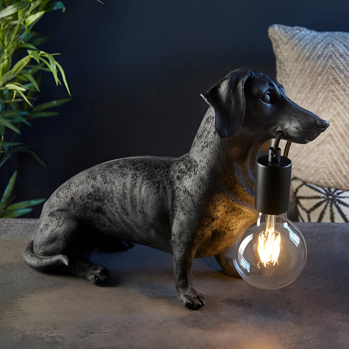 Bevworth Table Lamp in black resin, shaped as a seated dachshund holding an E27 filament bulb in its mouth, set against a dark room backdrop.
