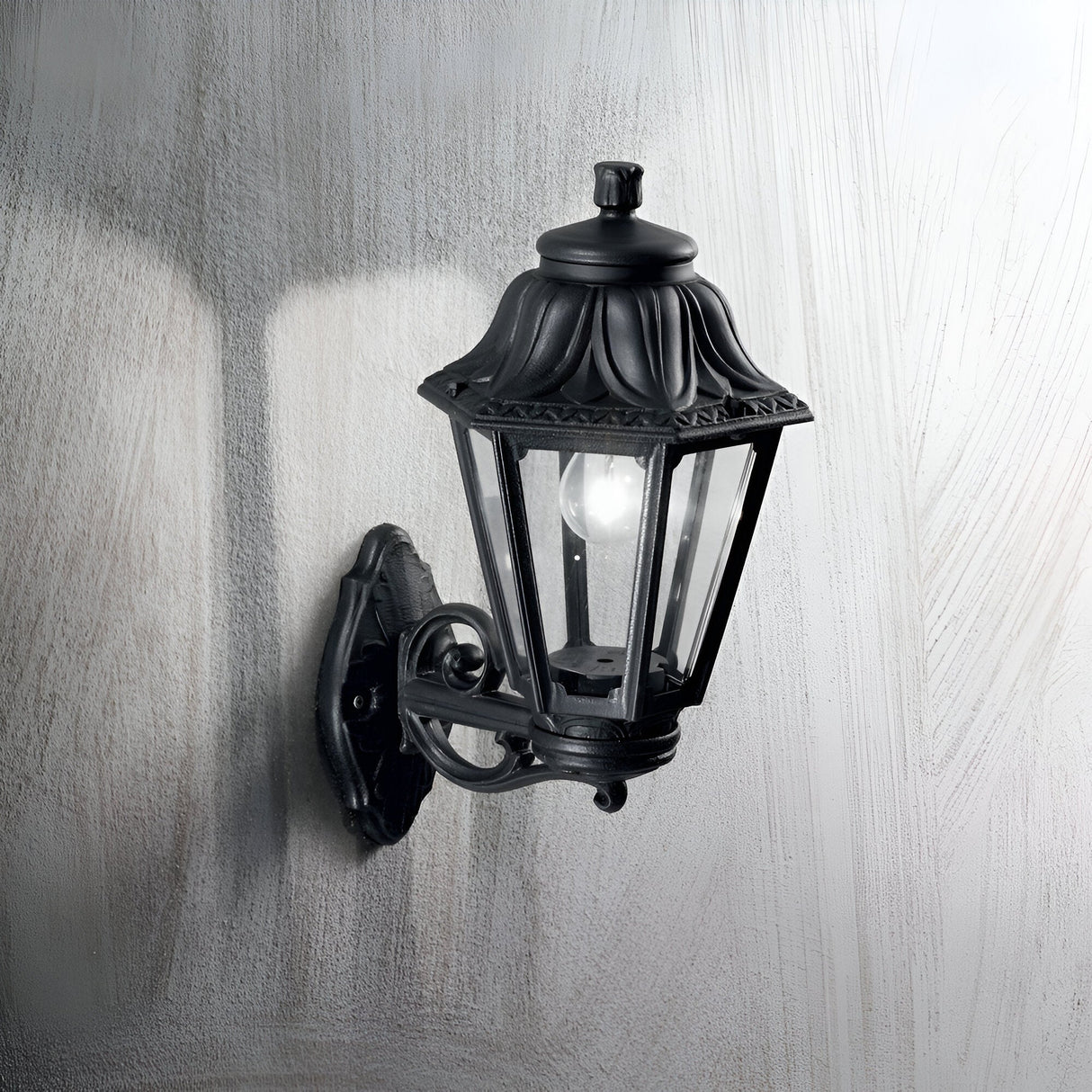 The Gaze Large Outdoor Wall Light in black, featuring a lantern-style design with glass panels and a visible bulb, is mounted on a light-colored textured wall. Crafted from UV-treated resin, it perfectly balances vintage charm with modern durability.