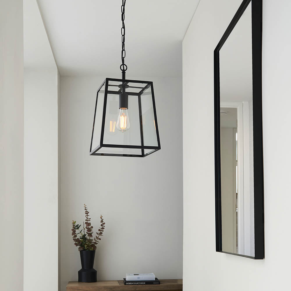 Brenton Lantern Pendant in black, a tapered box-frame ceiling light with clear glass panels and visible filament bulb, hung from a chain in a hallway.